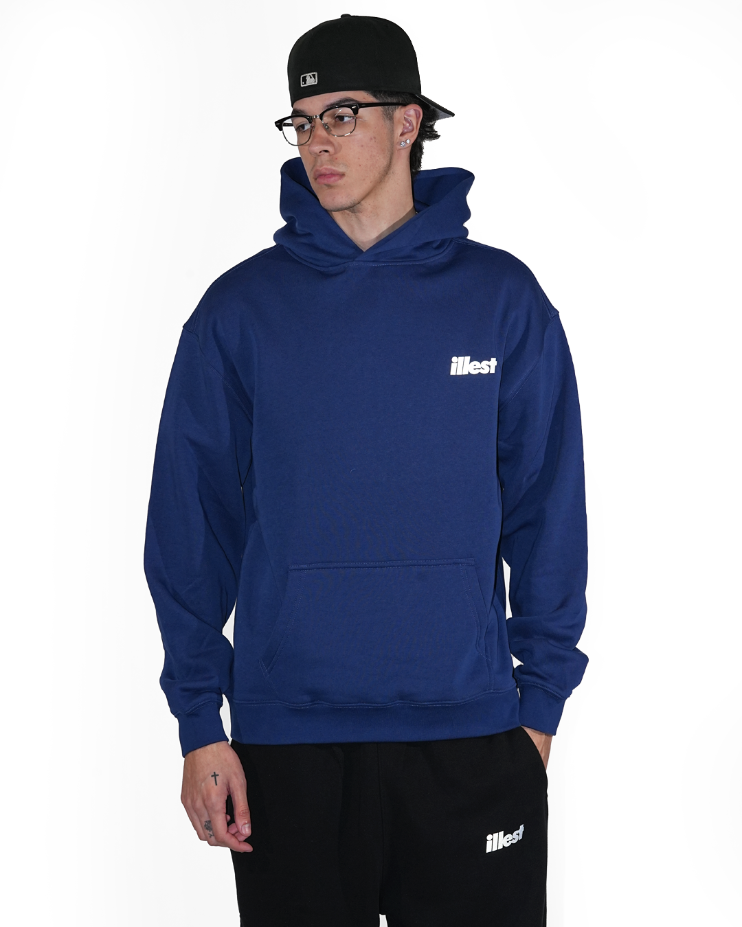 Male model wearing a blue Illest Prime Essentials hoodie with a front pocket, paired with black pants and a black cap.