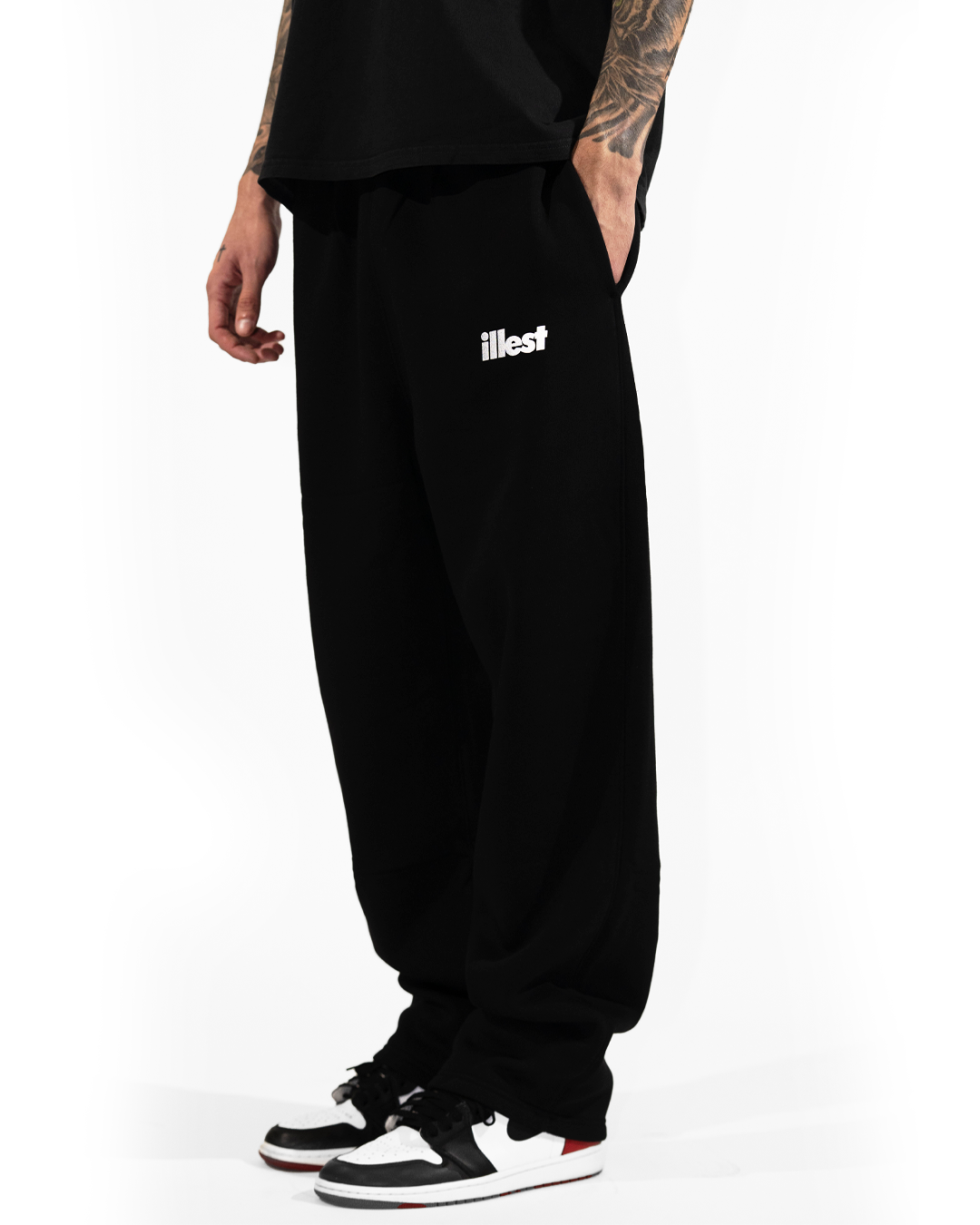 Black loose fit sweatpants with white Illest logo on the left thigh, paired with sneakers, worn by a person with tattoos.
