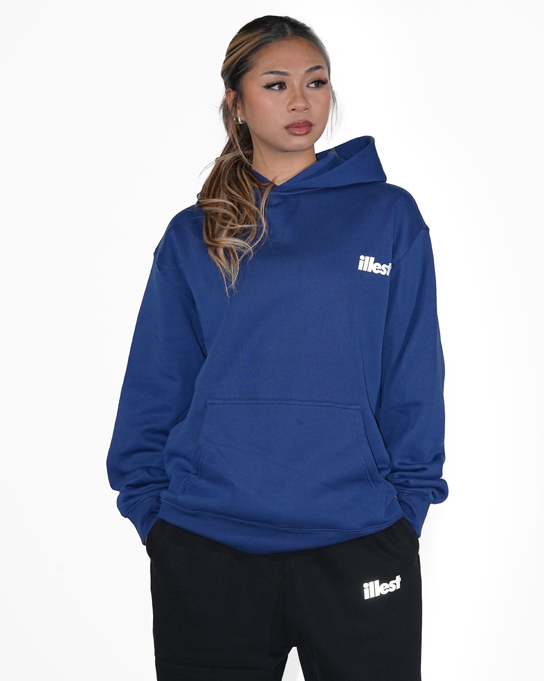 Woman wearing a blue Illest Prime Essentials hoodie with a front pocket, paired with black pants featuring the Illest logo.