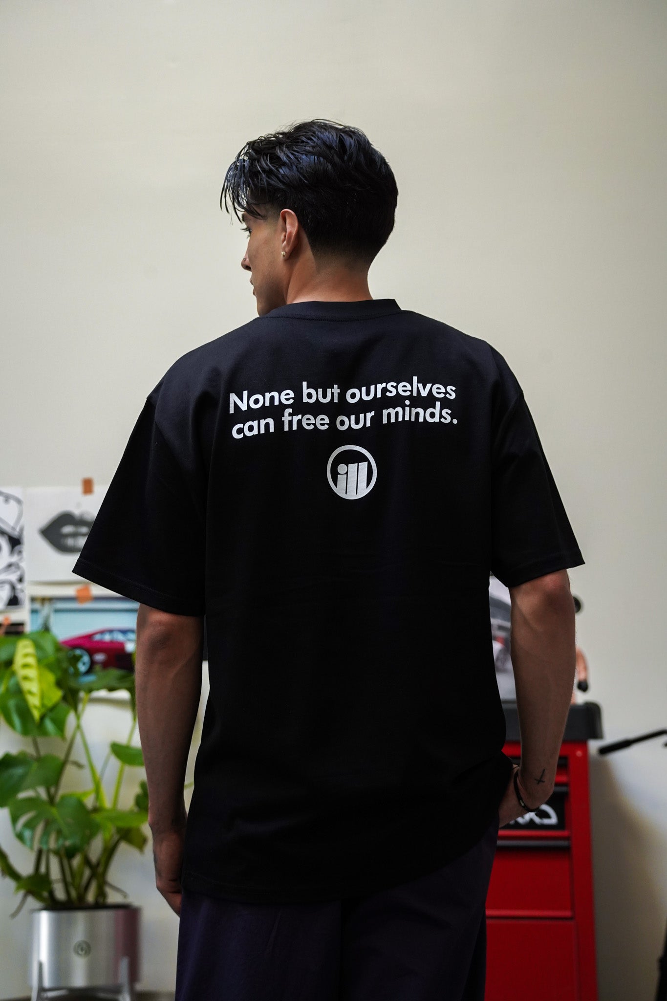 Back view of a person wearing a black Illest Bold Logo T-shirt with white text and logo, standing indoors near a plant and red toolbox.