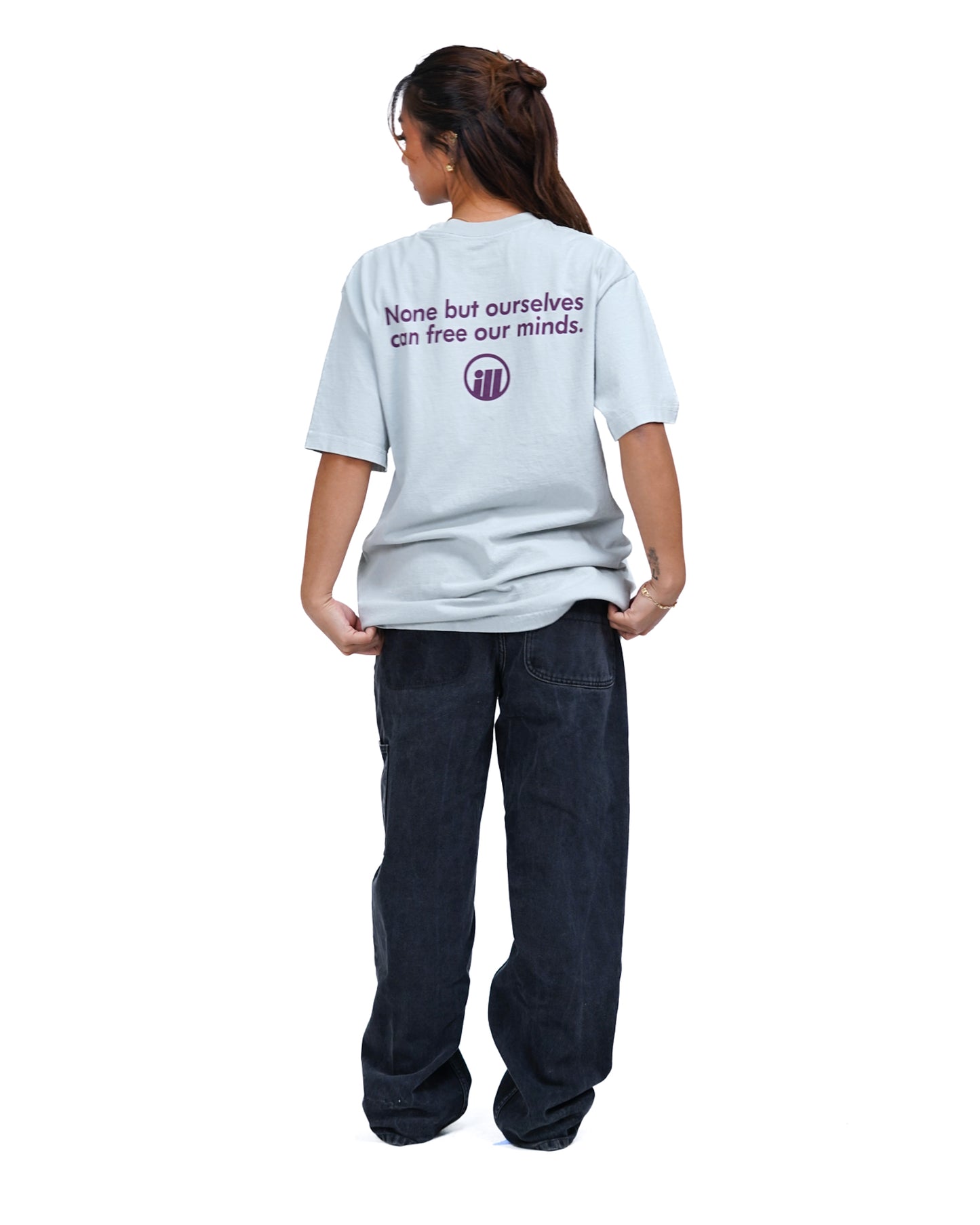 Back view of a woman wearing a light gray Illest Bold Logo Foam T-shirt with purple text and logo, paired with loose black jeans.