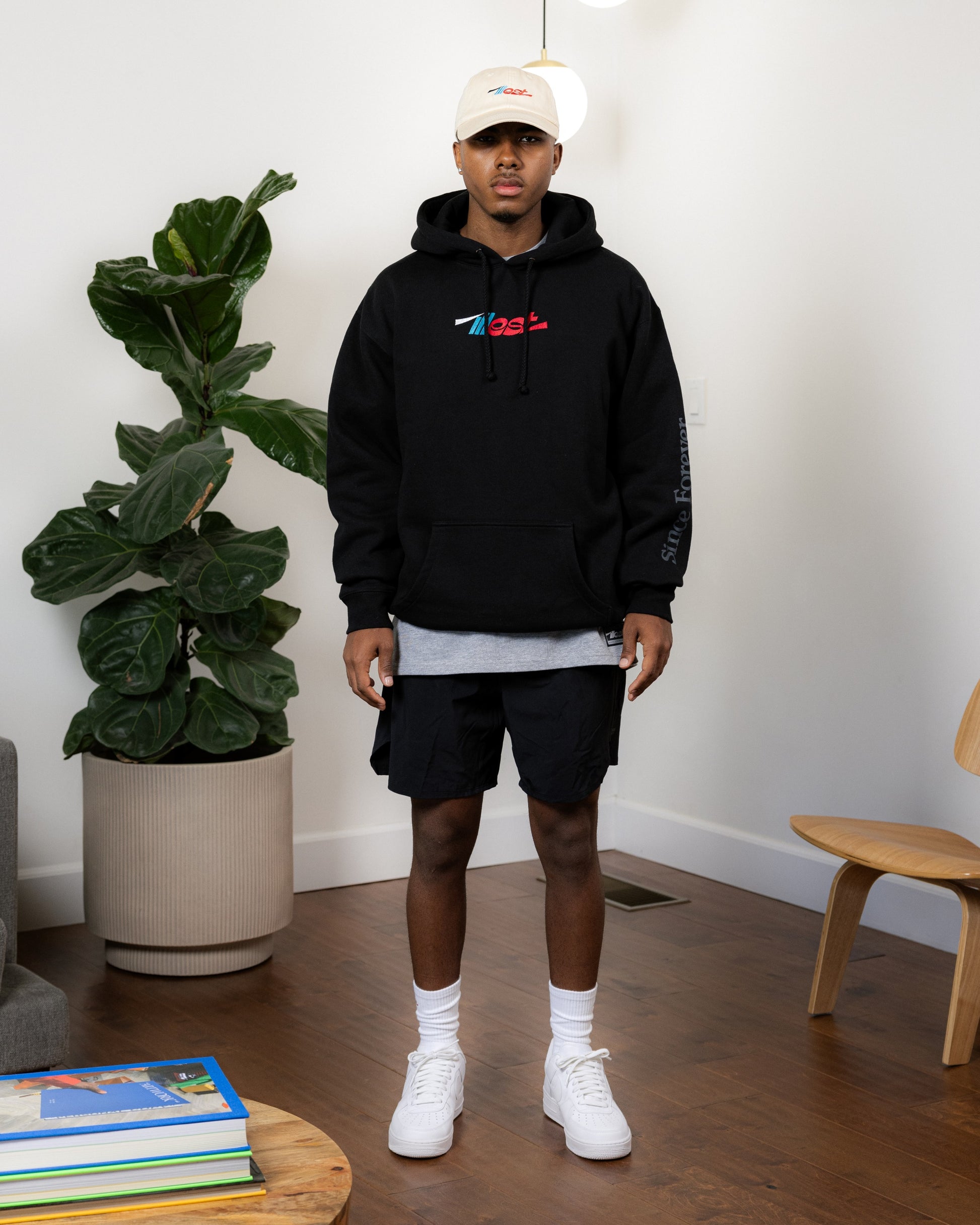 Young man wearing a black Illest Essentials Hoodie with colorful logo, paired with black shorts, white sneakers, and a beige cap.