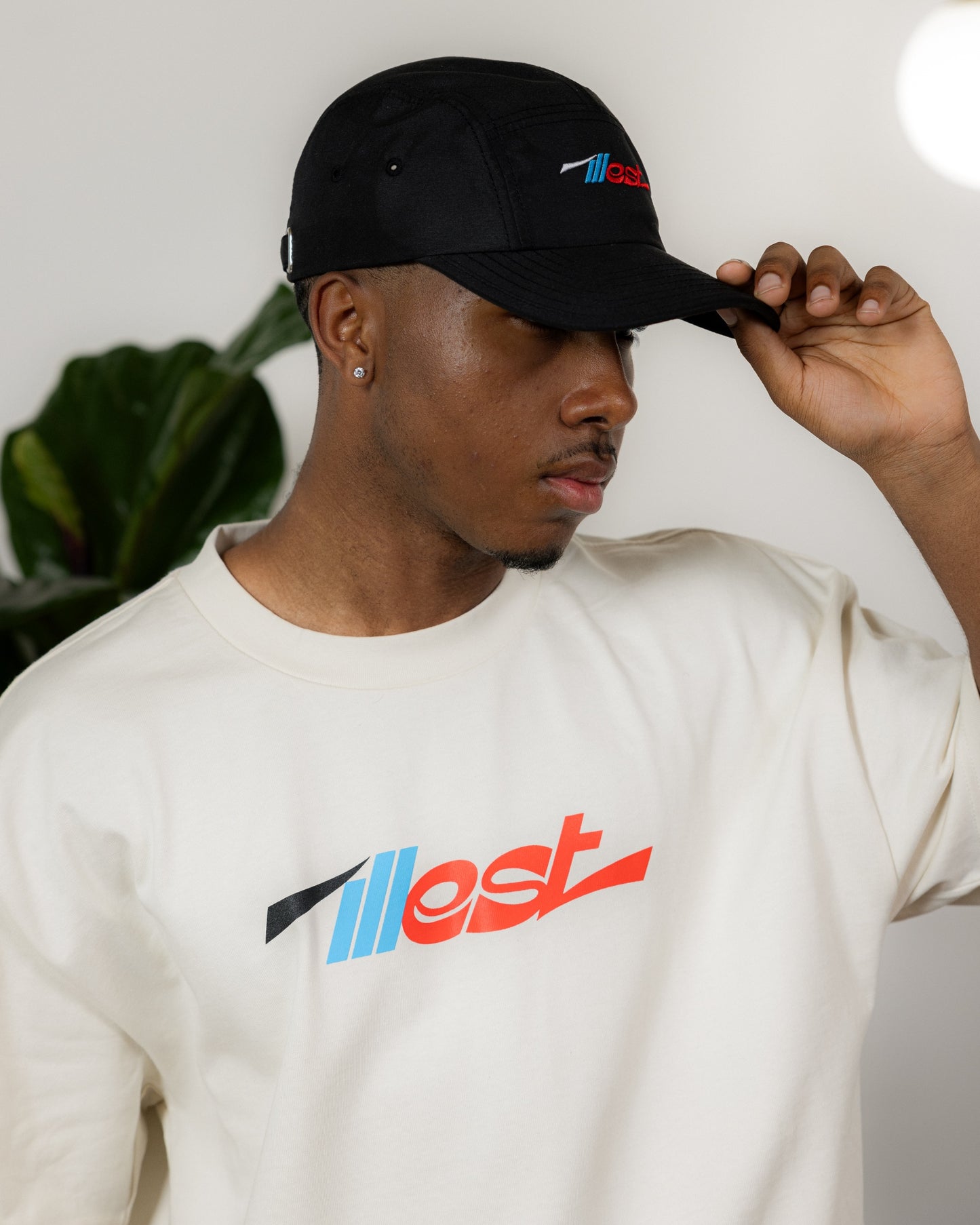 A man wears a black five-panel cap with colorful embroidered logo, paired with a cream T-shirt featuring a matching logo.