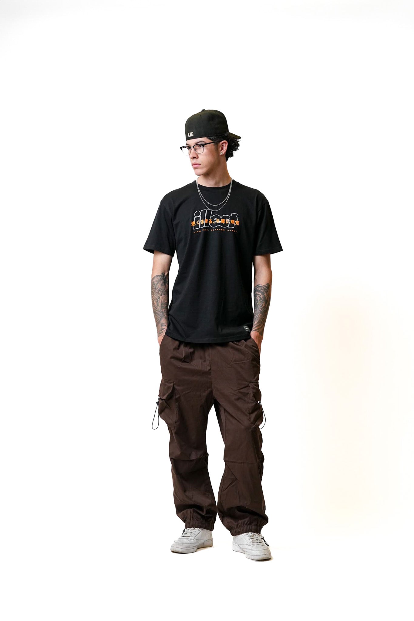 Young man wearing a black Illest Live Fast T-shirt, brown cargo pants, white sneakers, glasses, and a black cap, standing against a white background.