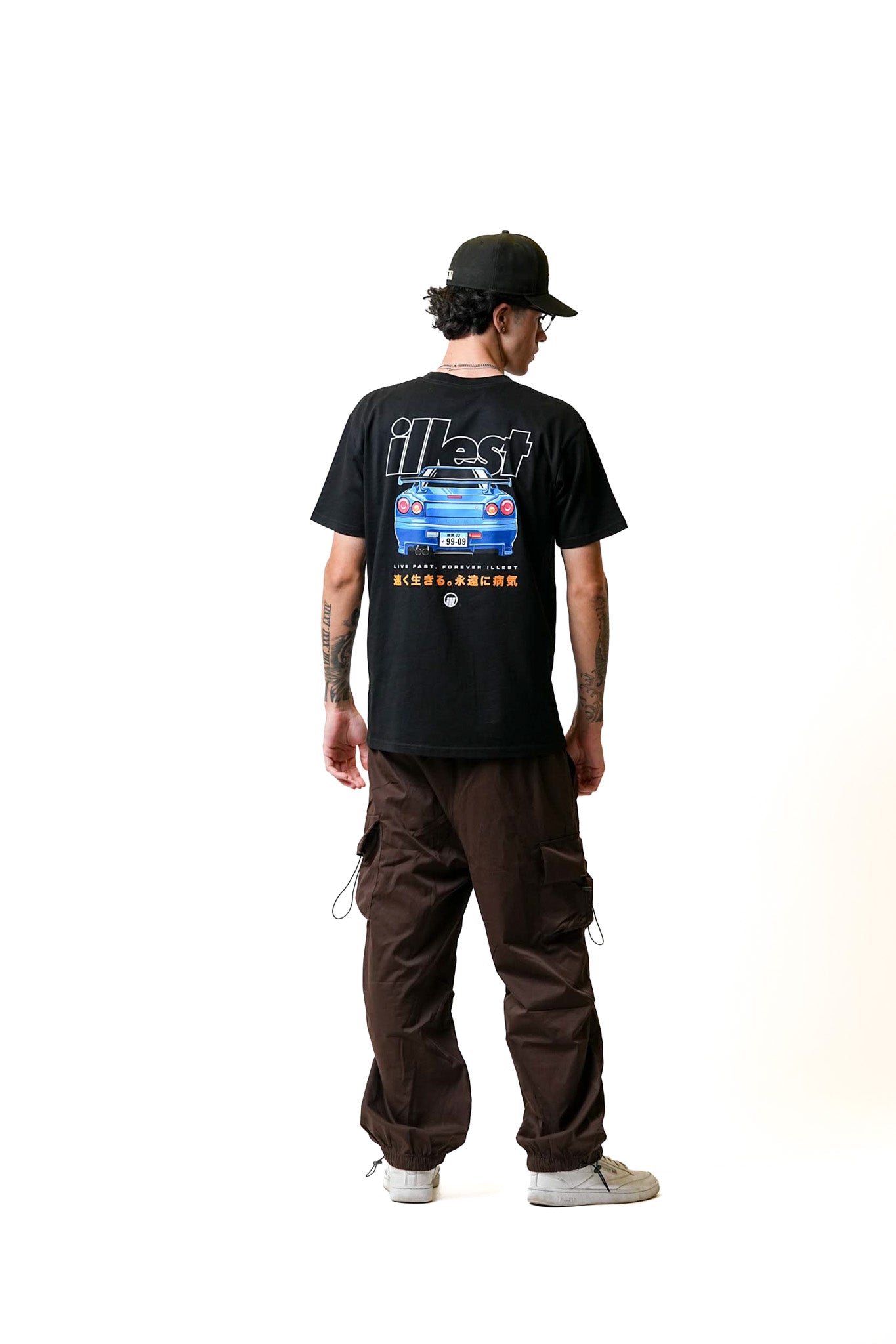 Back view of a person wearing a black Illest Live Fast T-shirt with a graphic of a blue car and Japanese characters, paired with brown cargo pants and a black cap.