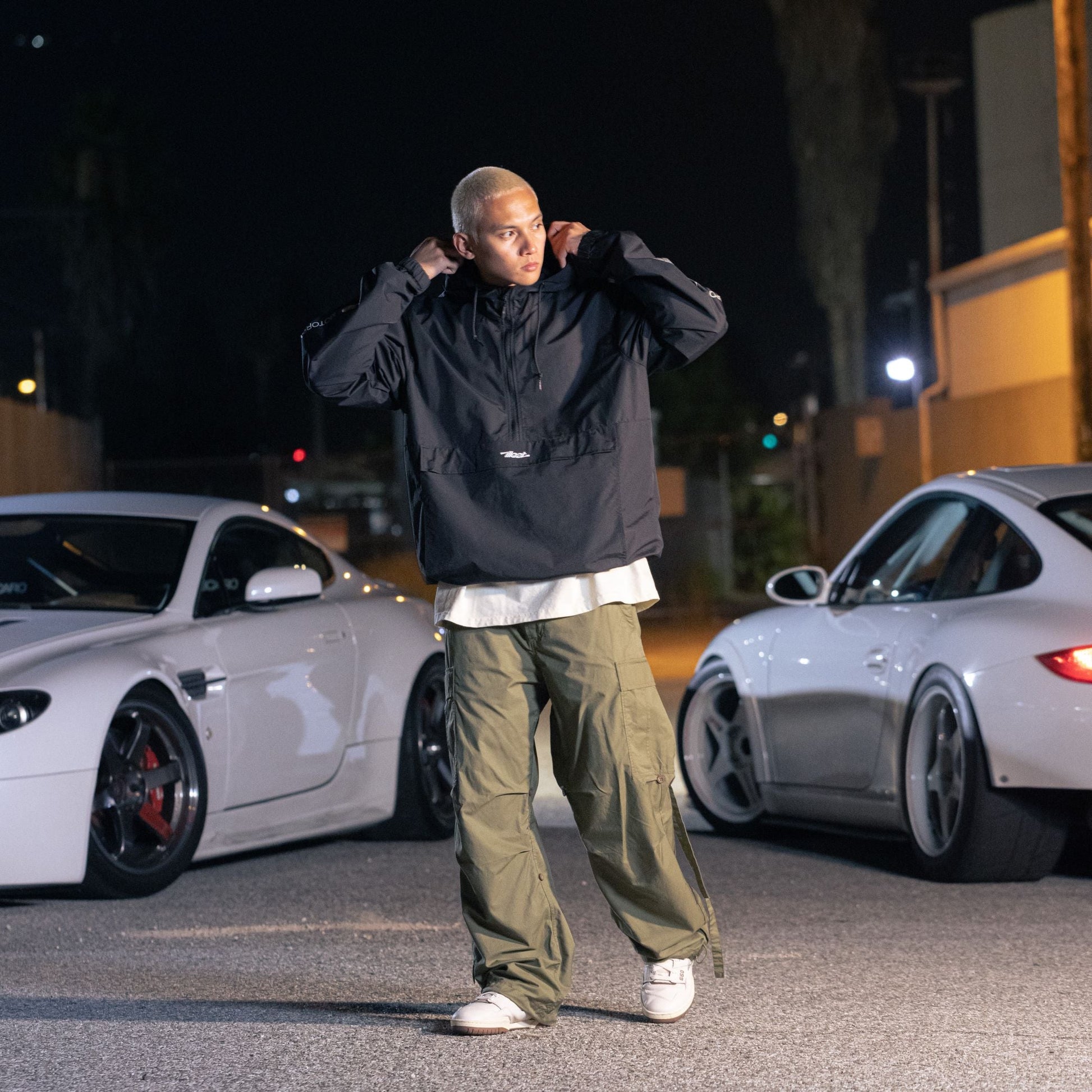 Person wearing a black Since Forever Anorak jacket, beige cargo pants, and white sneakers standing outdoors at night near white sports cars.
