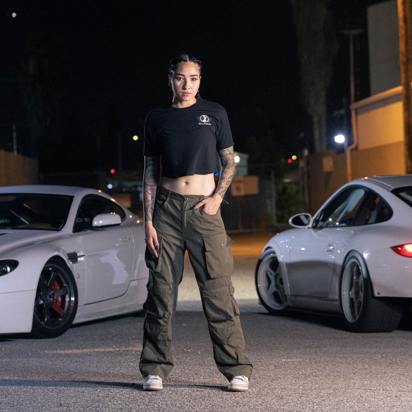 A woman stands outdoors at night wearing a black cropped T-shirt with "Since Forever" logo, paired with cargo pants and sneakers.