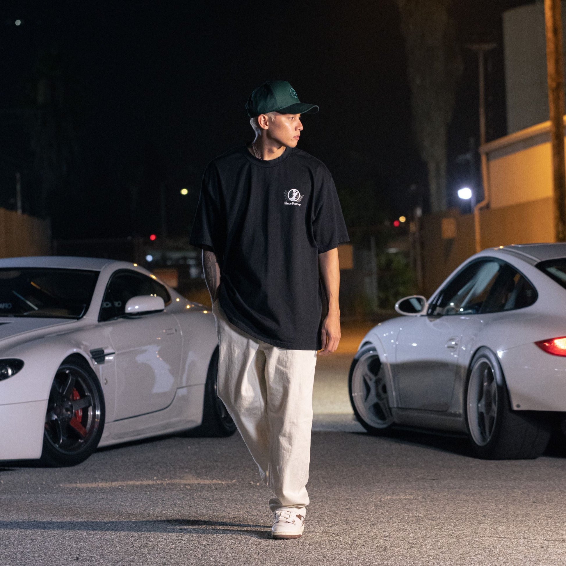 A young man wearing a black Since Forever Black Tee, beige pants, and a black cap stands next to white sports cars on a city street at night.