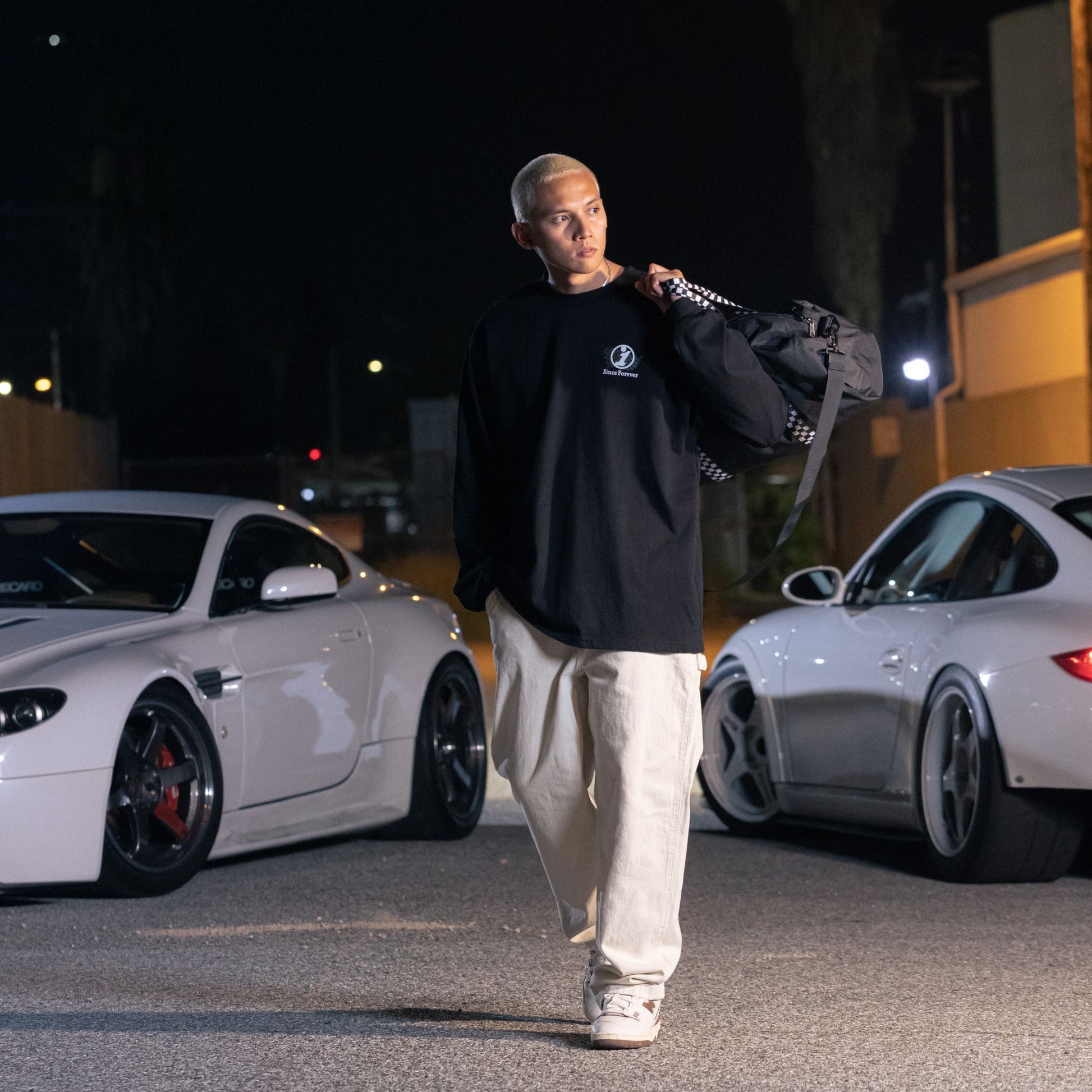 A person with short blonde hair wearing a black Since Forever Black LS Tee, beige pants, and white sneakers, standing next to white sports cars at night.