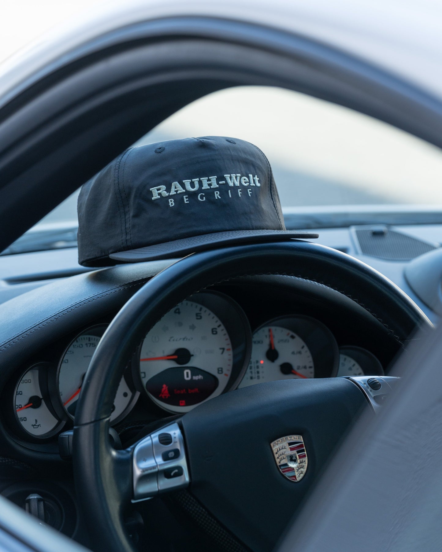 Black cap with "Rauh-Welt Begriffe" embroidered, placed on the dashboard of a Porsche sports car interior.