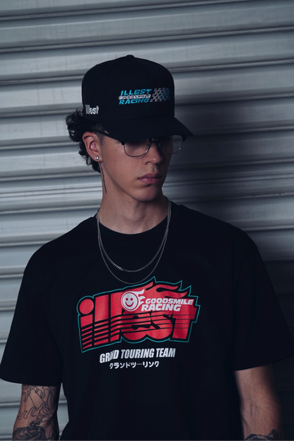 A young man wears a black ILLEST x GOODSMILE GT TEE with racing graphics and Japanese text, accessorized with a black cap and glasses.
