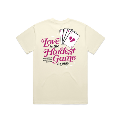 Cream-colored T-shirt with a graphic of playing cards and the phrase Love is the Hardest Game to Play, with additional text about heartbreaks and heartaches.
