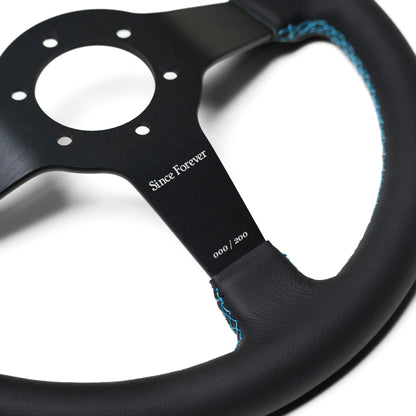Close-up of the Since Forever steering wheel with black leather grip and blue stitching, featuring a metal center with engraved branding.