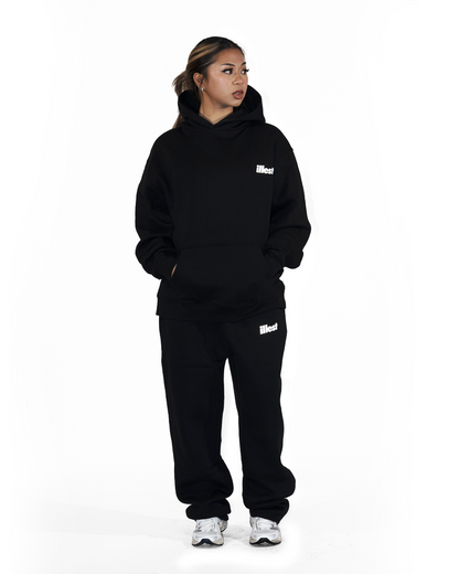 Woman wearing black Illest Prime Essentials Loose Fit Sweatpants and hoodie, standing with hands in pockets against a white background.