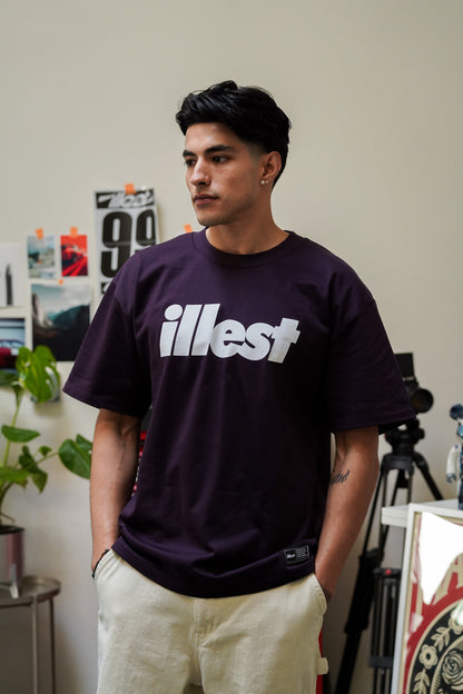 Young man wearing a dark purple Illest Bold Logo Plum Tee with white text, standing indoors with hands in beige pants.