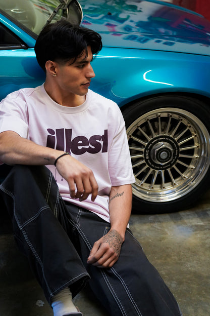 A young man with dark hair wearing a white Illest Bold Logo Orchid Tee, sitting on the floor next to a blue car with gold rims.