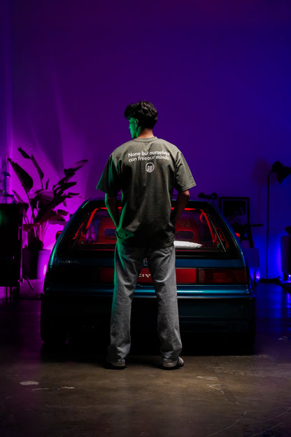 A person stands with their back to the camera in front of a car, wearing a gray Illest Bold Logo Cypress Tee in a dimly lit room with colorful purple and green lighting.