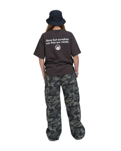 Back view of a person wearing a black Illest Bold Logo Mulch Tee with white text and a logo, paired with camouflage cargo pants and a black bucket hat.