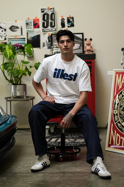 Young man sitting on a stool wearing a white Illest Bold Logo White Tee, dark jeans, and white sneakers in a creative room.