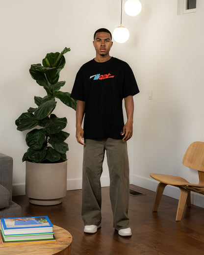 Young man wearing a black Illest Essentials T-shirt with colorful logo, beige pants, and white sneakers, standing indoors near a large potted plant.