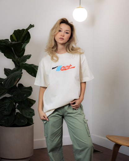 Woman wearing a white Illest Essentials Tee with colorful logo, paired with green cargo pants, standing indoors near a large potted plant.