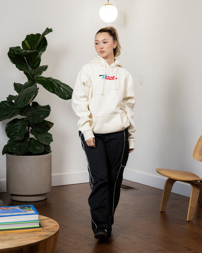 Woman wearing a cream Illest Essentials Hoodie with a colorful logo, paired with black track pants with white piping, indoors.