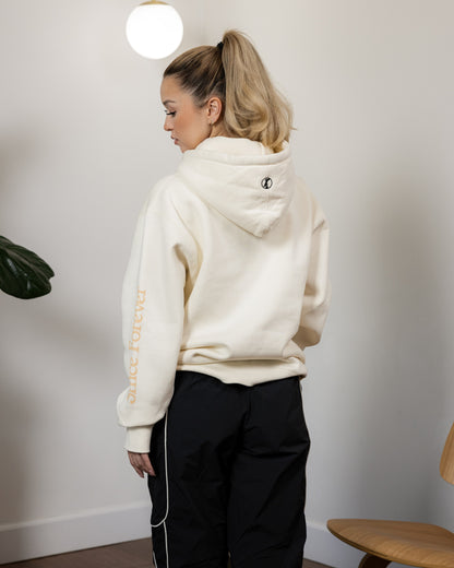 A woman wearing an off-white Illest Essentials Hoodie with "Once Forever" text on the sleeve, standing indoors with a neutral background.