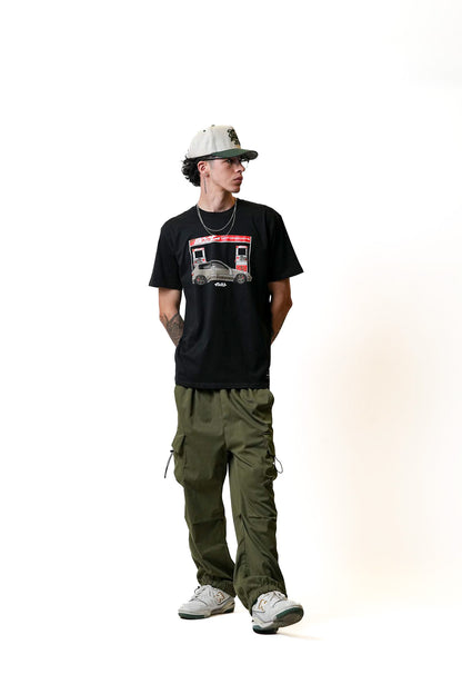 A person wearing an Illest Filling Station T-shirt, olive cargo pants, white sneakers, and a beige cap with a black brim.