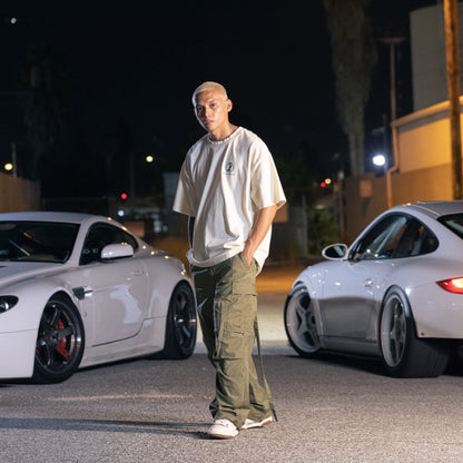 A young man with platinum blonde hair stands in a parking lot at night, wearing a white Since Forever Butter Tee and cargo pants, with two white sports cars behind him.