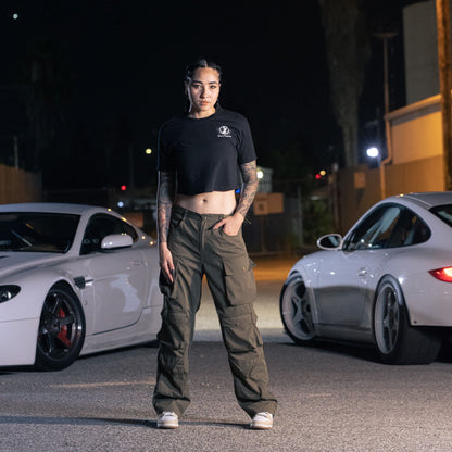 A woman stands outdoors at night wearing a black cropped T-shirt with "Since Forever" logo, paired with cargo pants and sneakers.