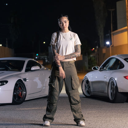 A woman wearing a white Since Forever Butter Crop Top and cargo pants standing in front of two white sports cars at night.
