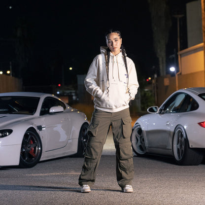 Young woman wearing a white Since Forever Dashboard Hoodie and cargo pants standing outdoors at night near two white sports cars.