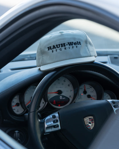 A beige dad cap with "RAUH-Welt BEGRIFF" embroidered in black, resting on the dashboard of a Porsche sports car.