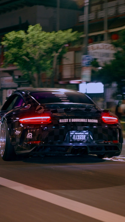 Black sports car with ILLEST x GOODSMILE RACING branding on the rear window and license plate, driving at night on an urban street.